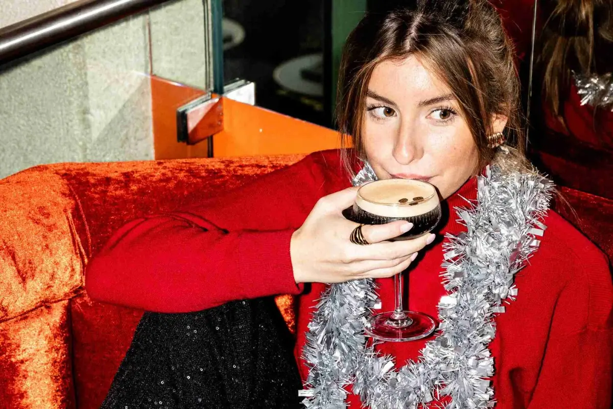 Cliente en pull rouge et guirlande de Noël, un cocktail à la main, sur une banquette en velours de Madame Claude